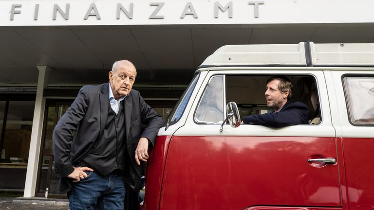 "Wilsberg - Datenleck": Wilsberg (Leonard Lansink) steht vor dem Finanzamtsgebäude und lehnt sich an einen kleinen Camper-Bus, in welchem Ekki (Oliver Korittke) am Steuer sitzt und begeistert hinausschaut.