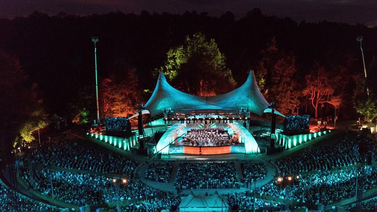 "Die Wiener Philharmoniker in der Waldbühne Berlin": Totale der Berliner Waldbühne bei Nacht, stimmungsvoll in Licht getaucht. Auf der Bühne spielt ein Orchester, im Vordergrund sind die vollbesetzten Publikumsränge.