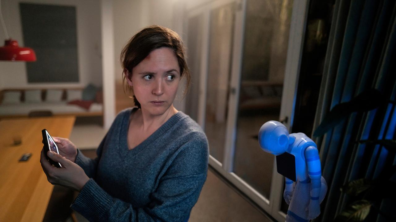 "Unsichtbarer Angreifer": Es ist dunkel im Haus und Emma (Emily Cox) ist offenbar allein. Auch Hausroboter Samy steht ausgeschaltet in der Ecke. Der Bildschirm auf Emmas Handy leuchtet, sie schaut besorgt um sich.