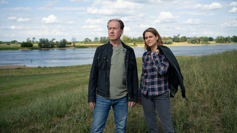  "Wendland - Stiller und der rote Faden": Jakob Stiller (Ulrich Noethen) und Kira Engelmann (Bettina Burchard) stehen zusammen auf einer Wiese am Ufer.