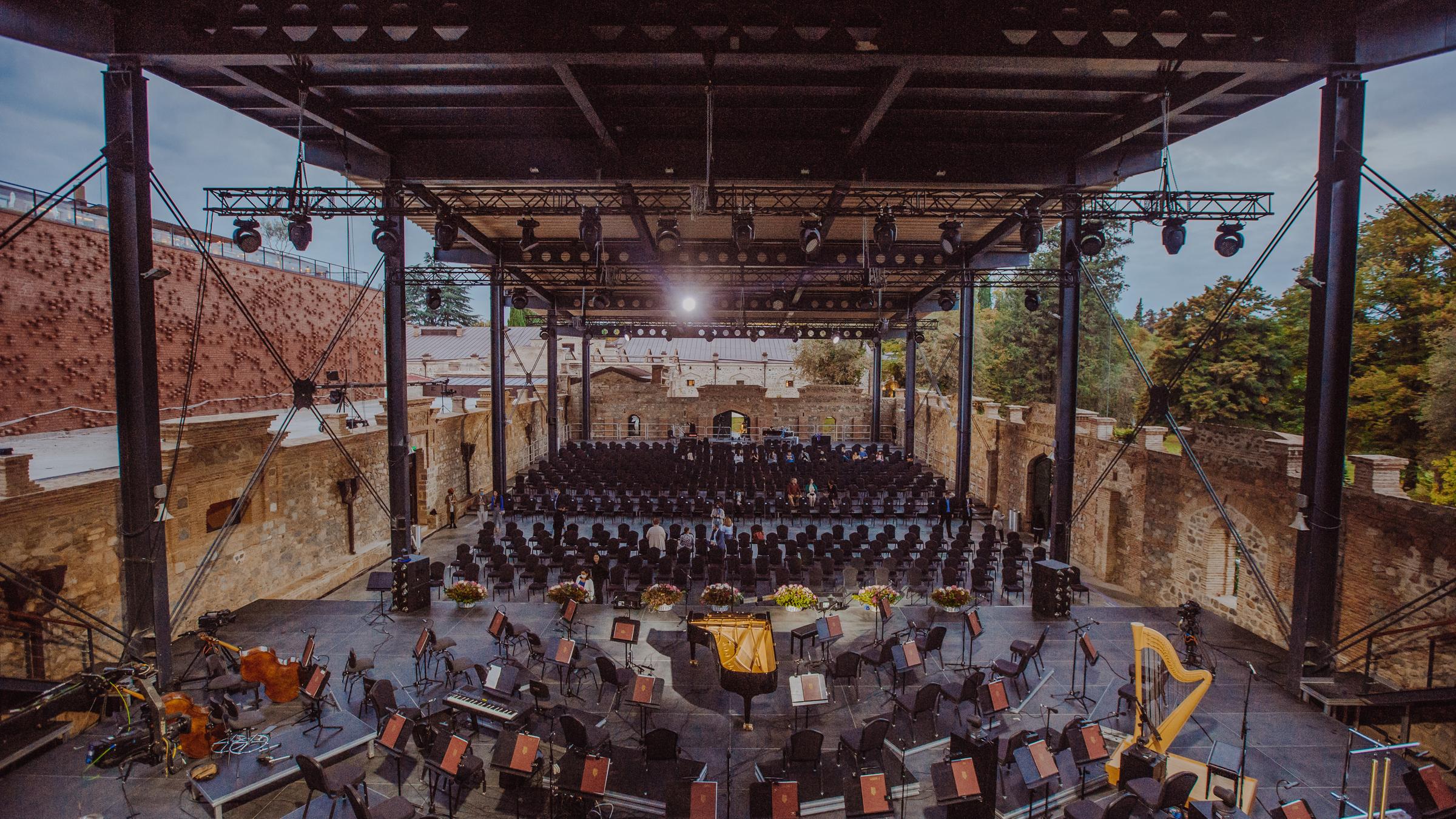 "Europakonzert 2024 aus Tsinandali in Georgien": Blick von schräg oben in ein Amphitheater im Stil eines Theaterraums mit Bestuhlung für Publikum: Im Vordergrund ist eine Bühne mit Stühlen für die Orchestermusiker, es stehen Notenpulte. Dahinter stehen Sitzreihen für das Publikum. Das Amphitheater ist zum Teil überdacht.