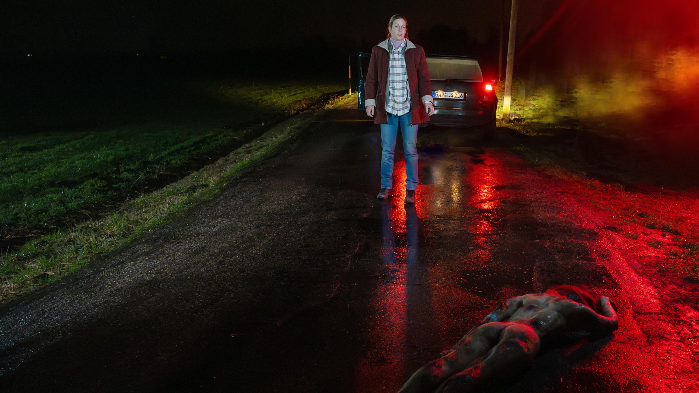 "XY gelöst - Tote ohne Gesicht": Eine Frau steht bei Nacht auf einer verlassenen Straße. Sie trägt eine Jeans und einen braunen Mantel und steht einige Meter vor ihrem geparkten Auto. Vor ihr liegt ein nackter Körper mit dem Gesicht zum Boden.