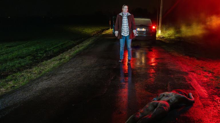 "XY gelöst - Tote ohne Gesicht": Eine Frau steht bei Nacht auf einer verlassenen Straße. Sie trägt eine Jeans und einen braunen Mantel und steht einige Meter vor ihrem geparkten Auto. Vor ihr liegt ein nackter Körper mit dem Gesicht zum Boden.