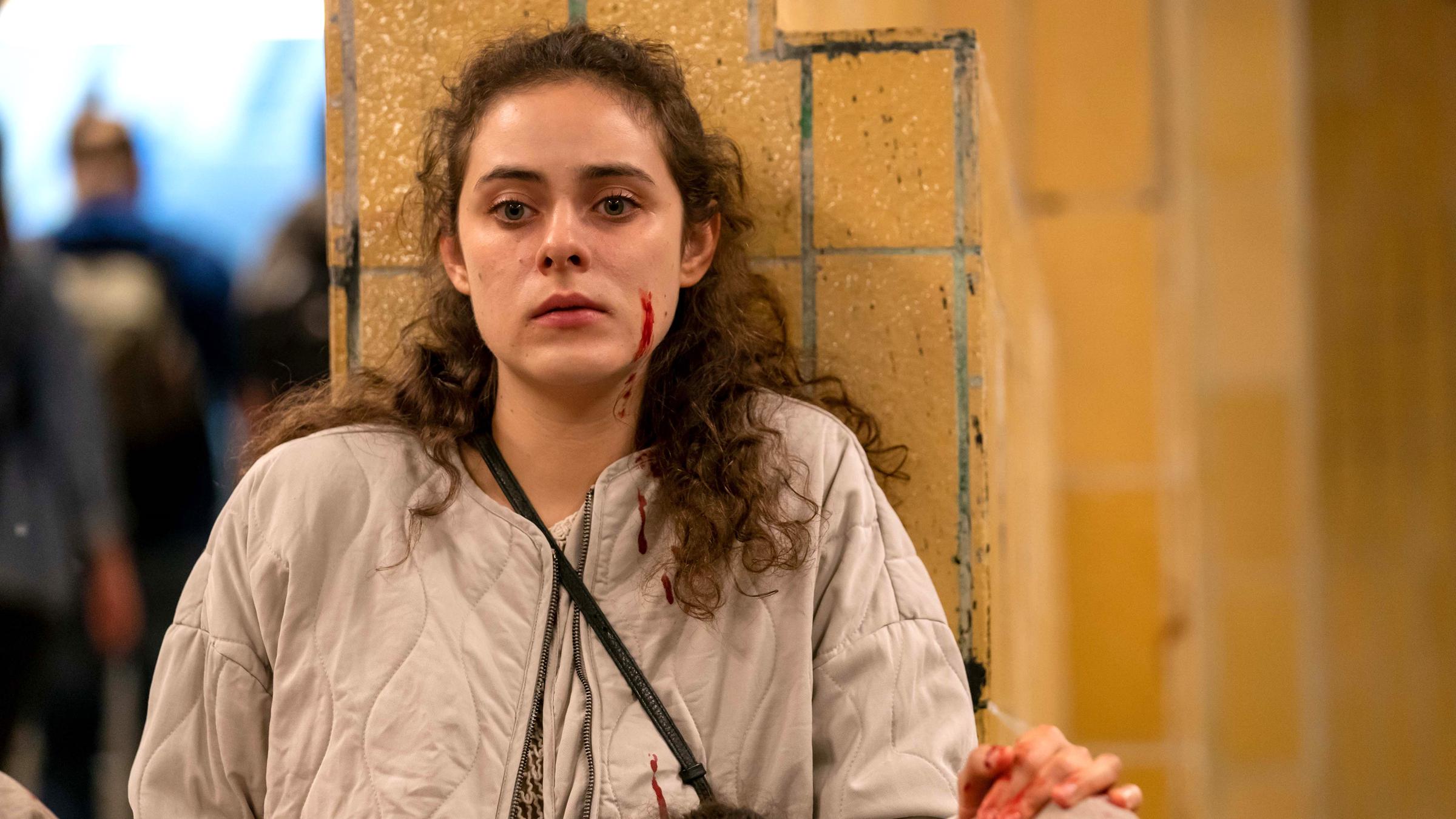 "Das Quartett - Das Schweigen": Julia (Emma Floßmann) sitzt mit dem Rücken gegen eine Mauer gelehnt. In ihrem Schoß liegt die tote Anna (ebenfalls Emma Floßmann). Julia schaut leer nach vorne. Auf ihren Händen, ihrer Jacke und in ihrem Gesicht befindet sich Blut.