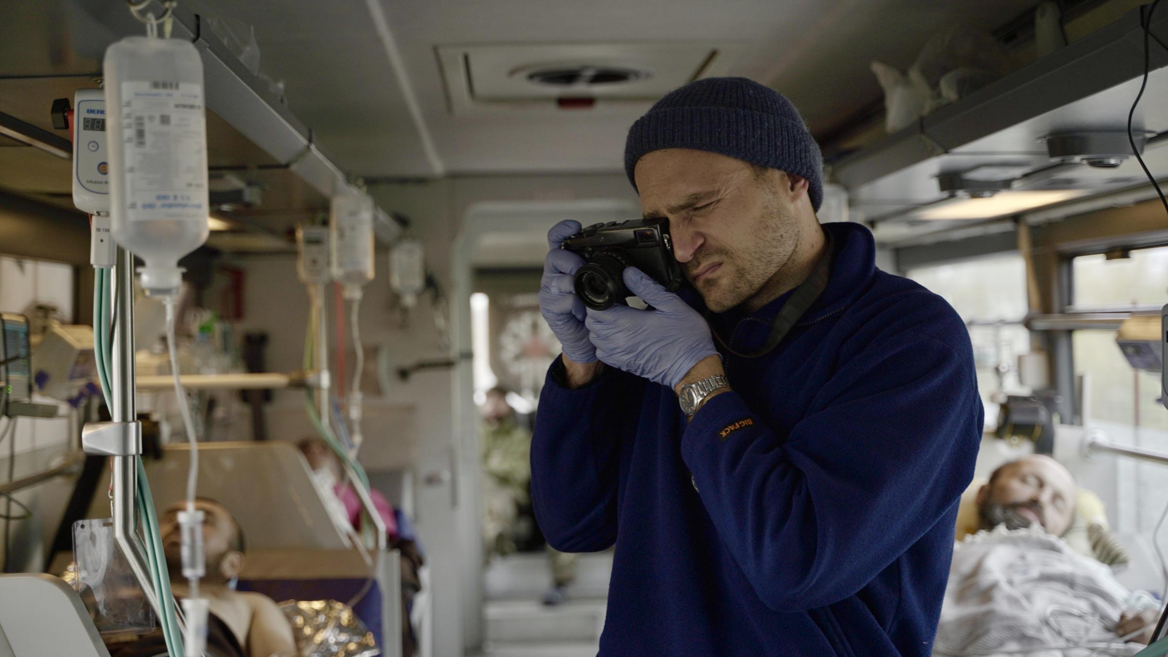 "37°: Gefährlicher Einsatz - Journalist*innen berichten aus der Ukraine": In einem Bus, der mit Liegen und medizinischem Equipment speziell für Krankentransporte ausgestattet wurde, steht Vincent Haiges und fotografiert, ein Auge zugekniffen, das andere durch den Sucher seiner Kamera schauend. Er trägt einen dicken, blauen Pullover, Wollmütze und medizinische Handschuhe. Neben ihm hängt ein Infusionsbeutel der einen Soldaten versorgt. Weiter hinten liegen weitere verwundete Soldaten.