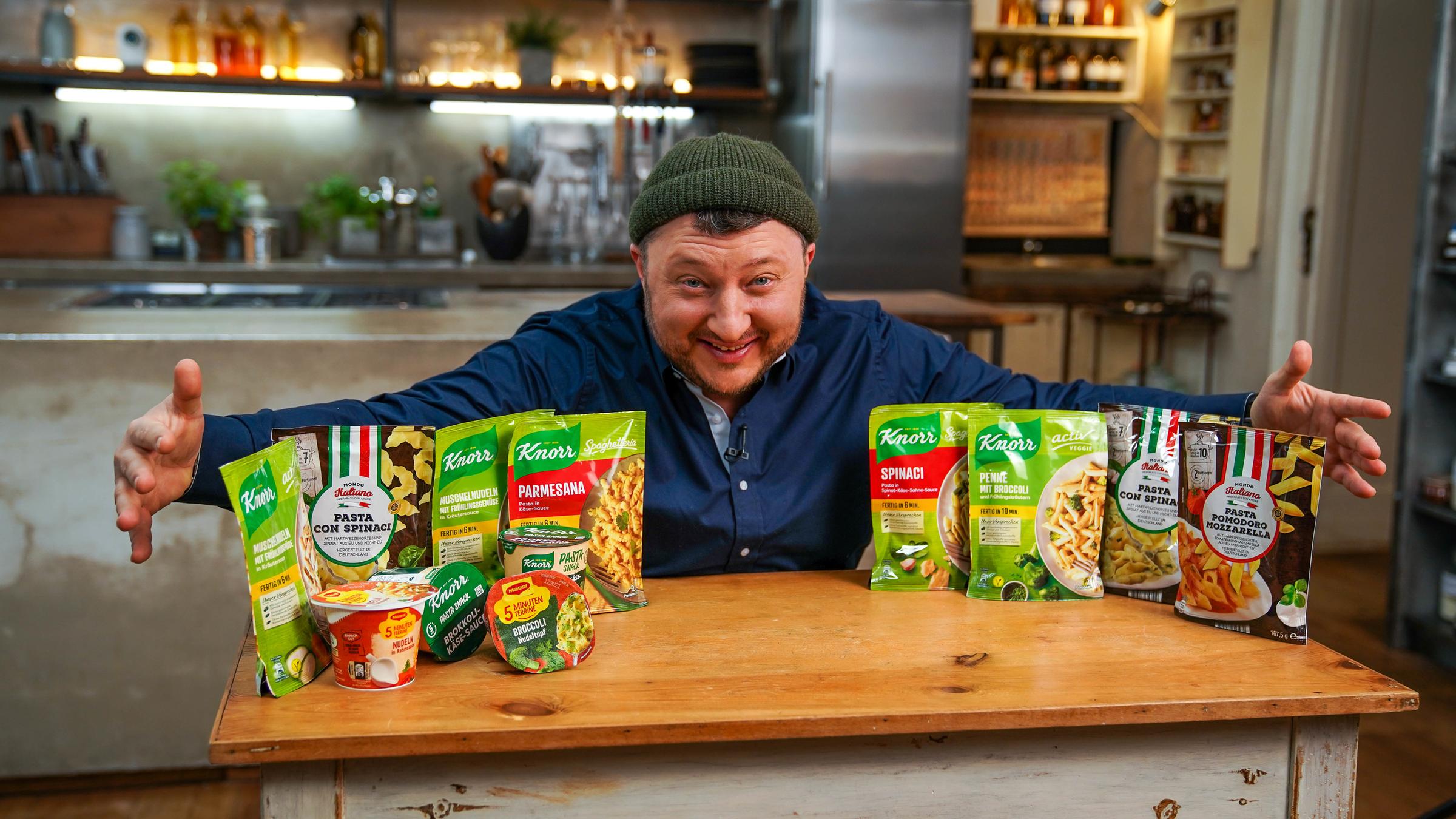 "besseresser: Leere Gesundheitsversprechen - Sebastian Lege packt aus": Sebastian Lege steht hinter einem Tisch auf dem Tütennudelpackungen stehen.
