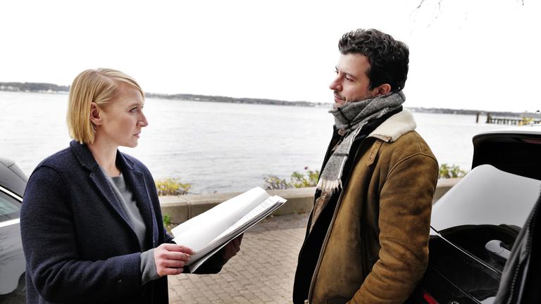 "Solo für Weiss - Tödliche Wahl": Nora Weiss (Anna Maria Mühe) und ihr Kollege Ben Salawi (Camill Jammal) stehen auf der Straße am Ufer neben ihren Autos. Nora Weiss (Anna Maria Mühe) hält Unterlagen in der Hand.