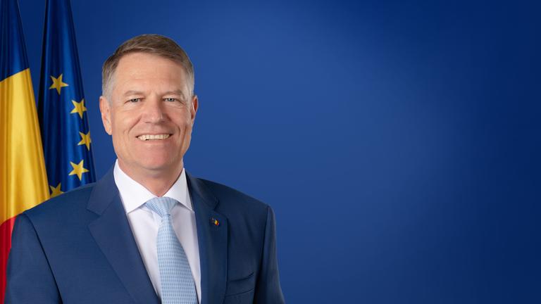 "Gedenkstunde zum Volkstrauertag": Der rumänische Präsident Klaus Werner Iohannis steht lächelnd vor einem blauen Hintergrund. Hinter ihm sind die Flaggen Rumäniens und Europas zu sehen.