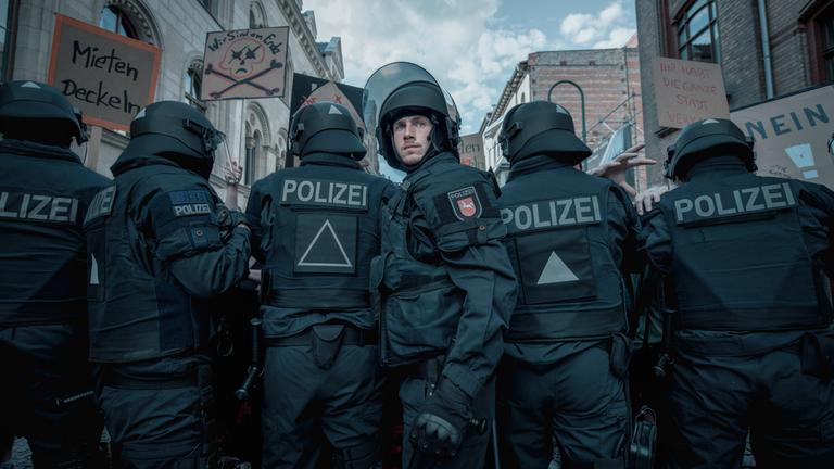 "Allein zwischen den Fronten": Jan Vogt (Justus Johanssen) steht vor einem mit schwarzer Einsatzkleidung und Helmen bestückten Einsatztrupp der Polizei und blickt nach vorne in die Kamera. Über den Personen sind Demonstrationsschilder und einige Hausfassaden zu erkennen.