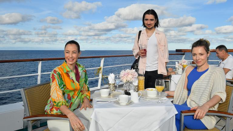 "Das Traumschiff: Argentinien": Auf dem Schiffsdeck: Glücklich lächelnd sitzen Cora Bruns (Friederike Linke) und die Yogalehrerin (Alessandra Meyer-Wölden) am gedeckten Frühstückstisch. Tobias Bender (Ivo Kortlang) steht am Tisch und hält ein gefülltes Glas in den Händen.