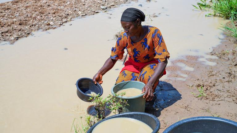 "planet e.: Klimawandel und Gender - Warum die Krise Frauen stärker trifft": Eine Afrikanerin kniet an einem Wasservorkommen und befüllt mehrere Eimer mit Wasser.