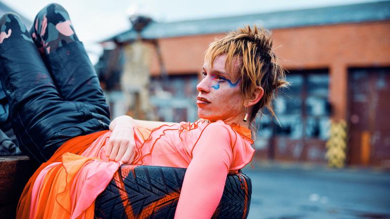 „KlassiXS – die großen Dramen in jungen Kurzfilmen - Splitter": Das Bild zeigt eine Person, die lässig auf einem Stapel Reifen liegt. Die Person trägt ein auffälliges, farbenfrohes Outfit mit einem leuchtend pinken Oberteil und einer orange-pinken, durchscheinenden Rock-ähnlichen Schicht. Die Hose ist dunkel mit einem Tarnmuster. Das Gesicht der Person ist markant geschminkt, mit blauen Akzenten um das rechte Auge und roten Lippen. Die Haare sind kurz und wild gestylt, in einem rötlich-blonden Ton. Im Hintergrund ist eine unscharfe städtische Umgebung mit Gebäuden und einem leicht bewölkten Himmel zu sehen.