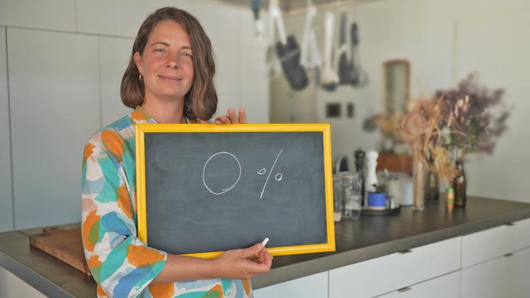 Eine junge Frau, Lisa Jaspers, steht in ihrer Küche und hält eine Tafel hoch, auf der mit Kreide 0 Prozent geschrieben steht.
