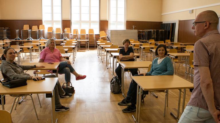 "Abi '89 - Neues Land und neue Chancen": In einer Aula sitzen vier Erwachsene an Tischen. Im Vordergrund steht ein Mann an einem Tisch gelehnt.