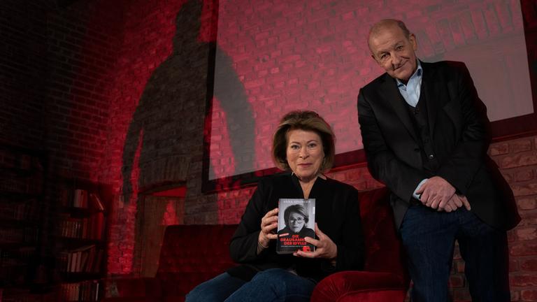 "Wilsberg - Blut geleckt": Anna Springer (Rita Russek) sitzt in einem großen roten Samtsessel und präsentiert vorsichtig lächelnd ihr "True Crime"-Buch "Die Grausamkeit der Idylle", welches sie in den Händen hält. Wilsberg (Leonard Lansink) sitzt auf der Lehne des Sessels und blickt ebenfalls lächelnd in die Kamera. Im Hintergrund erhebt sich der Schatten einer unbekannten Person an der Wand, der für Anna Springer und Wilsberg gefährlich wirkt.