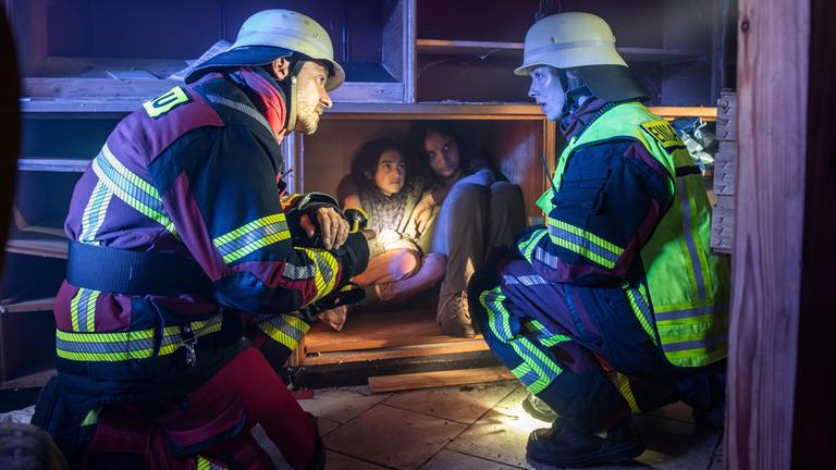 "Marie fängt Feuer - Aufbruch ins Ungewisse": Marie (Christine Eixenberger) kniet neben Peter Angerer (Stephan Luca) vor einem Holzschrank, in dem sich zwei Kinder versteckt halten. Beide tragen ihre Feuerwehruniform Helm und Taschenlampe und sehen sich an.