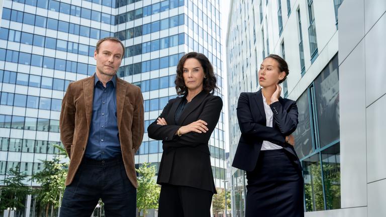 "Ein Fall für Conti - Spieler": Frank Stolpe (Mark Waschke), Anna Conti (Désirée Nosbusch) und Henry Mahn (Malaya Stern Takeda) stehen nebeneinander vor der gläsernen Fassade moderner Gebäude.