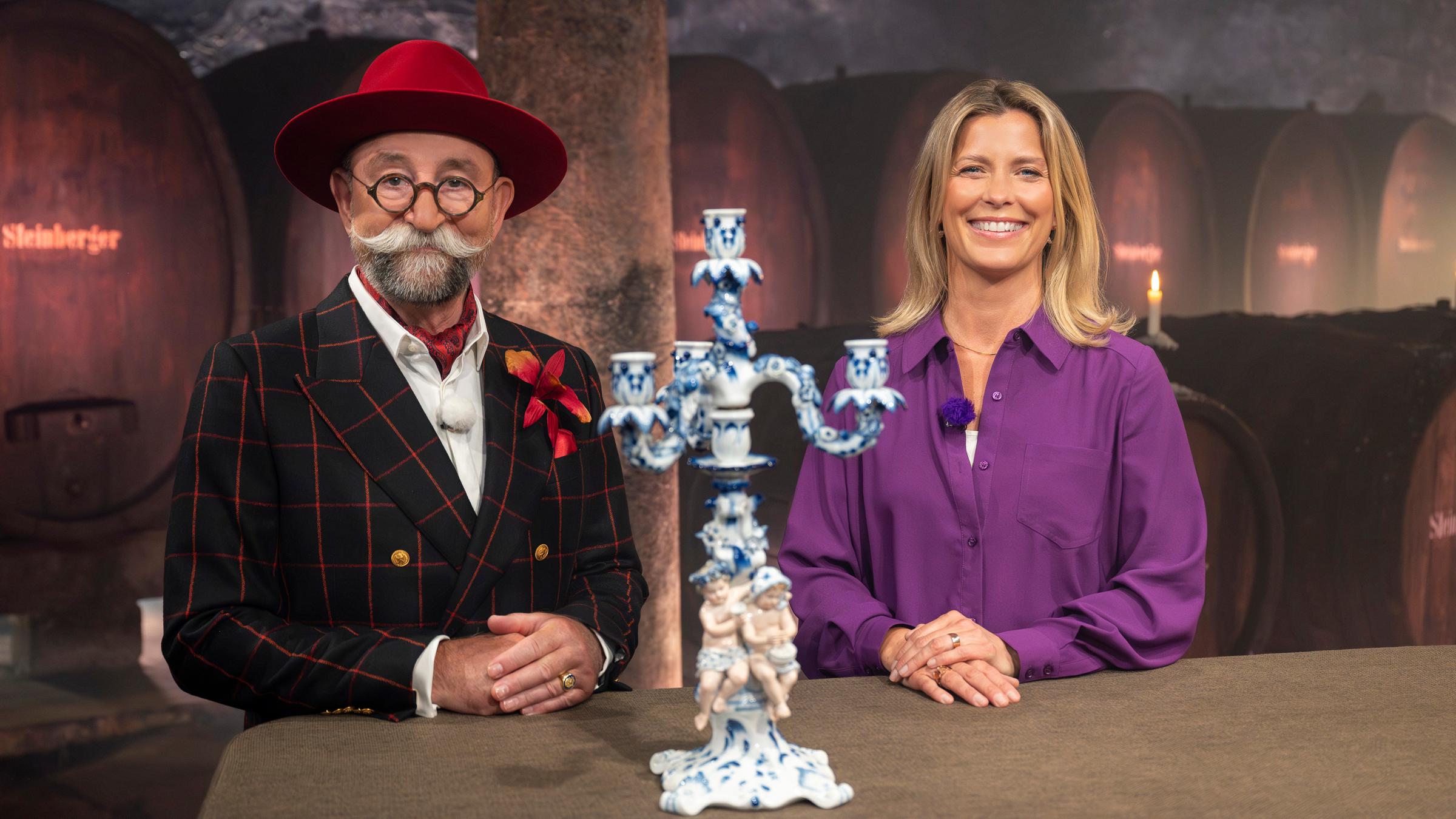 "Bares für Rares - Deutschlands größte Trödelshow": Horst Lichter und Valerie Niehaus stehen hinter dem Expertisentisch und lächeln in die Kamera. Zwischen ihnen steht ein Porzellanleuchter. Im Hintergrund stehen Weinfässer in einem Gewölbekeller.