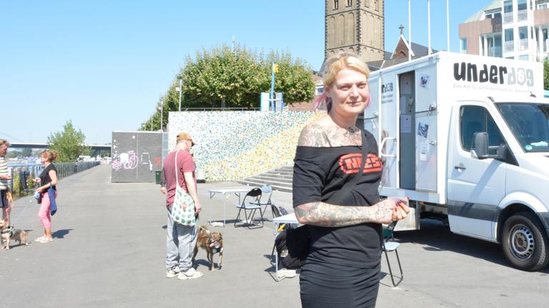 Bildbeschreibung: "Leben ist mehr! - Hilfe für Menschen und Hunde": An den Düsseldorfer Rheintreppen steht die Sozialarbeiterin Jana vor einem Wagen mit der Aufschrift "Underdog" und schaut lächelnd in die Kamera.