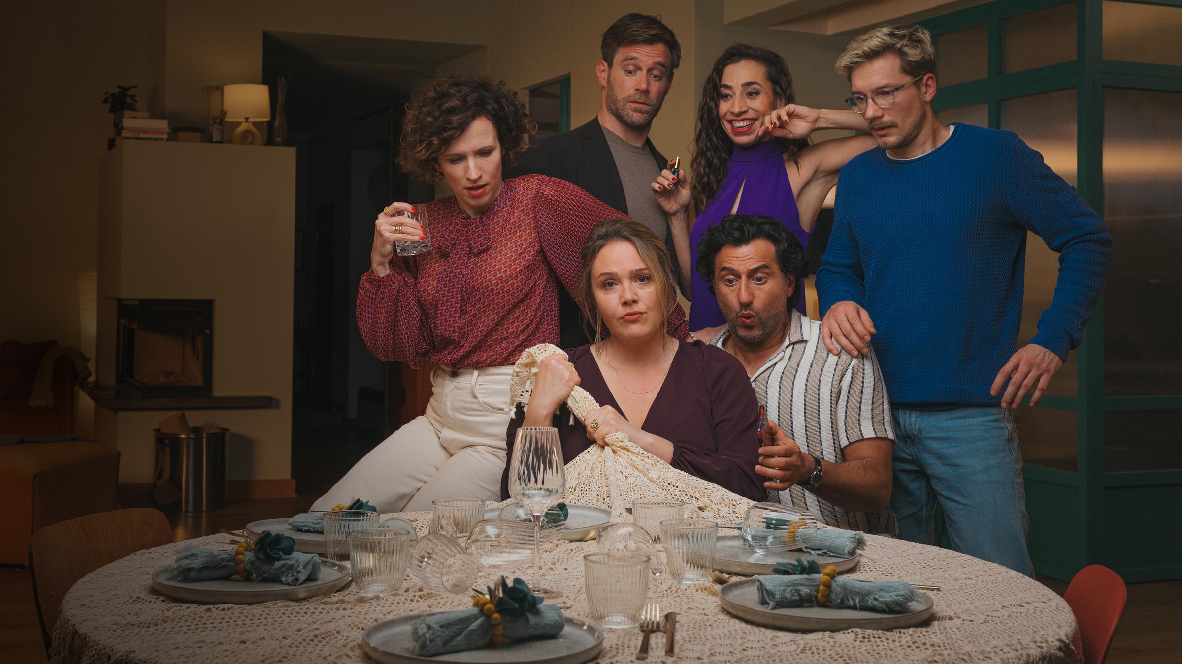 "Pärchenabend": Caro (Alwara Höfels) sitzt neben ihrem Mann Tarek (Serkan Kaya) an einem runden, gedeckten Tisch und zieht die gehäkelte Tischdecke vom Tisch.  Hinter den beiden stehen Anne (Adina Vetter), Phillip (Ken Duken), Nesrin (Carol Schuler) und Matze (Jacob Matschenz). Anne sitzt halb auf der rechten Stuhllehne von Caro und schaut zur Tischdecke herunter. Phillip steht hinter Anne und blickt mit hochgezogenen Augenbrauen zu Caro. Links von Phillip steht Nesrin, die mit großem Lächeln dabei zusieht, wie die Tischdecke vom Tisch gezogen wird. Ganz rechts steht Matze. Er macht einen nachdenklichen Gesichtsausdruck.