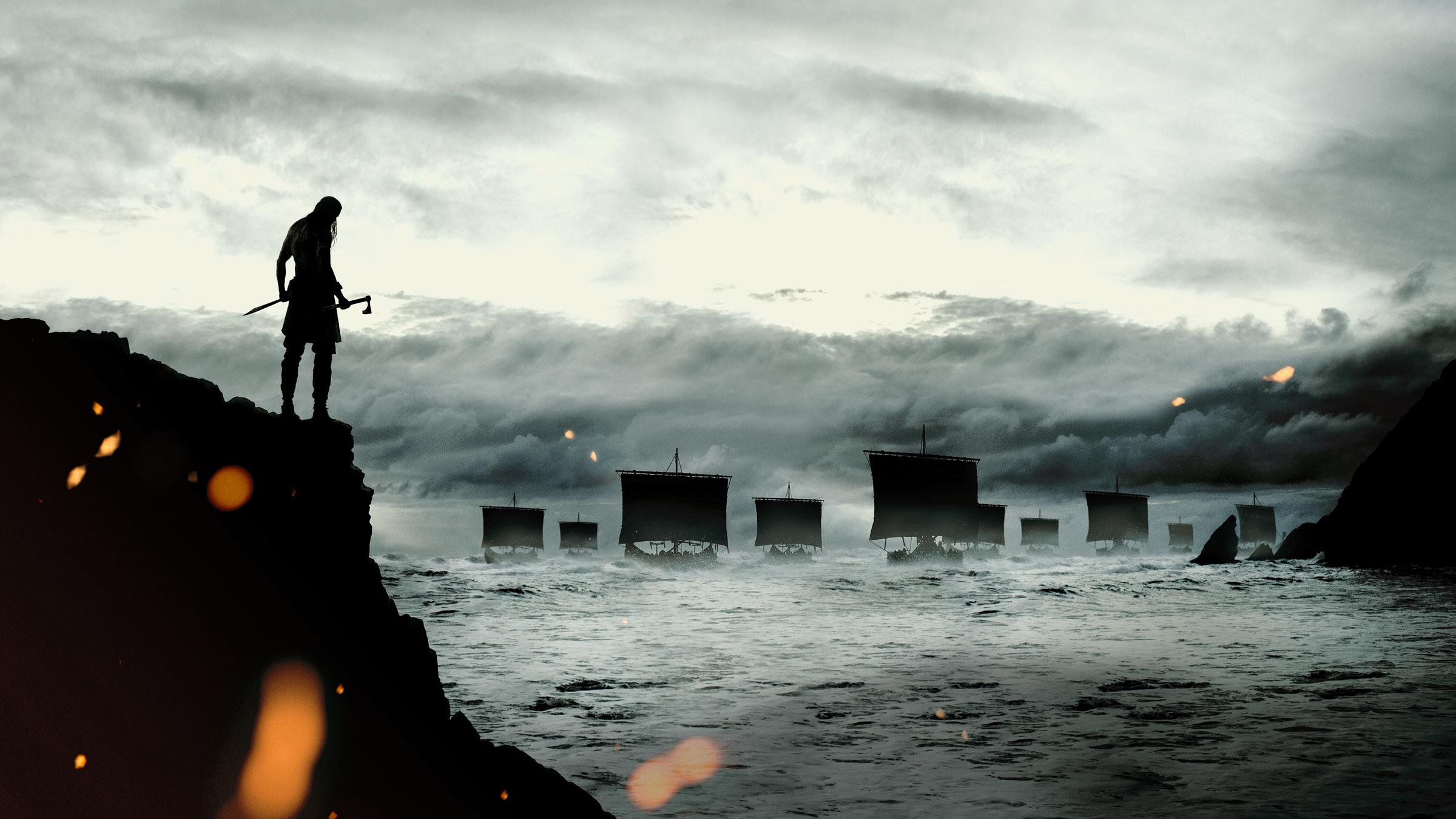  "The Northman": Totale: Die Silhouette von einem Mann mit Schwert und Beil ist auf einer Klippe über einem stürmischem Ozean sichtbar. Auf dem Meer fährt eine Armada von Kriegsschiffen. Der Himmel ist mit grauen Sturmwolken bedeckt.