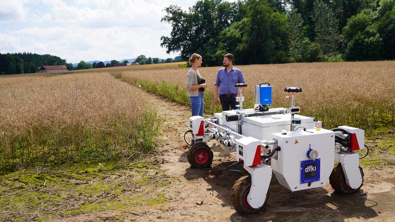 "plan b: Künstliche Intelligenz in der Landwirtschaft - Gegen den Hunger in der Welt": Judith Reese und Benjamin Kisliuk stehen mit einem weißen KI-Roboter auf einem Rapsfeld.