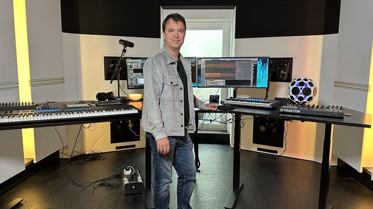 "Künstliche Musik": Mann mit kurzen Haaren in Jeans und Jeansjacke steht in Tonstudio, links ein Keyboard, in der Mitte und recht Computer und Monitore.