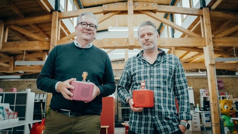 "WISO-Dokumentation: Zwischen Boom und Burnout - Startup-Gründer unter Druck": In einer Werkhalle: Marcus Stahl und Patric Faßbender halten in ihren Händen jeweils eine Toniebox und schauen lächelnd in die Kamera.
