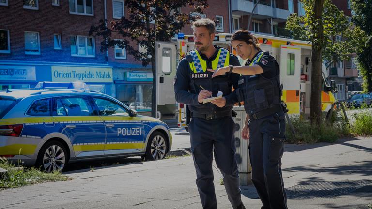 "Notruf Hafenkante - Neues Leben": Der Polizist Kris Freiberg (Marc Barthel) und seine Kollegin Isa Nowak (Lilli Hollunder) stehen dicht nebeneinander auf einem Bürgersteig. Isa weist mit ihrer Hand auf etwas vor ihnen. Kris hält einen Block und einen Stift in den Händen. Beide blicken in die Richtung, in die Isa zeigt. Seitlich neben ihnen am Straßenrand steht ein Streifenwagen und ein Notarztwagen.
