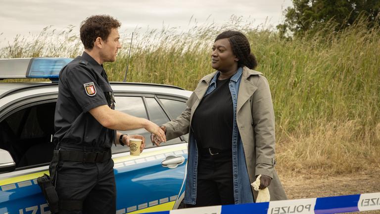 "Die Polizistin und die Sprache des Todes": Pieper Olsen (Artjom Gilz) und Gloria Acheampong (Thelma Buabeng) stehen neben einem Polizeiwagen und geben sich die Hand.