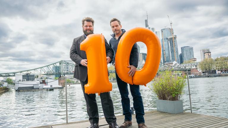 "Ein Fall für Zwei": Benjamin Hornberg (Antoine Monot) und Privatdetektiv Leo Oswald (Wanja Mues) stehen am Mainufer in Frankfurt und halten jeweils einen orangenen Luftballon in Form einer "1" bzw. einer "0", die zusammen die Zahl "10" bilden. 