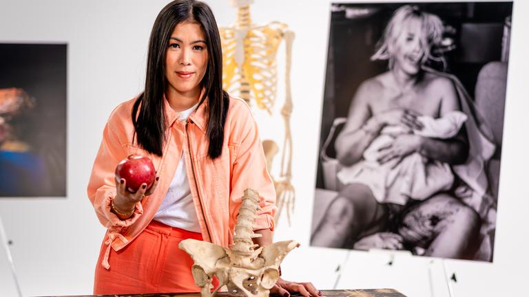 "Terra X: Unser Leben - wie es beginnt": Im Studio: Die Moderatorin Mai Thi Nguyen-Kim hält einen Granatapfel in den Händen und vor ihr steht ein Model des Beckens. Im Hintergrund ist ein Skelett und daneben das Foto einer Frau, die soeben ein Baby zur Welt gebracht hat.