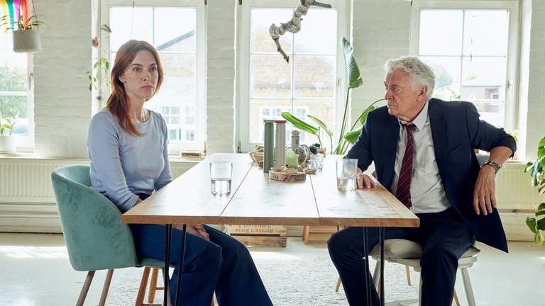 "Die Stille am Ende der Nacht": Liane Sievers (Kim Riedle) sitzt an einem langen Esstisch und starrt verschlossen nach vorne. Johannes Fischer (Henry Hübchen) sitzt an der anderen Seite des Tisches und schaut sie an.