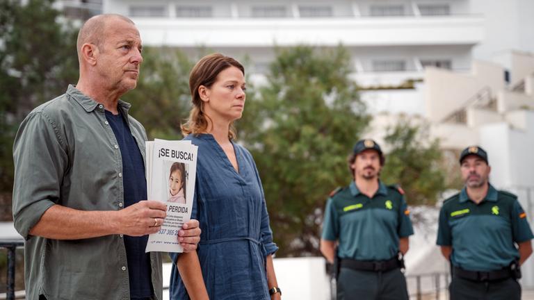"Lillys Verschwinden (1)": Robert Bischoff (Heino Ferch) und Anna Bischoff (Jessica Schwarz) stehen mit einem Plakat ihrer vermissten Tochter auf der Straße nebeneinander. Zwei Polizisten (Komparsen) stehen im Hintergrund.