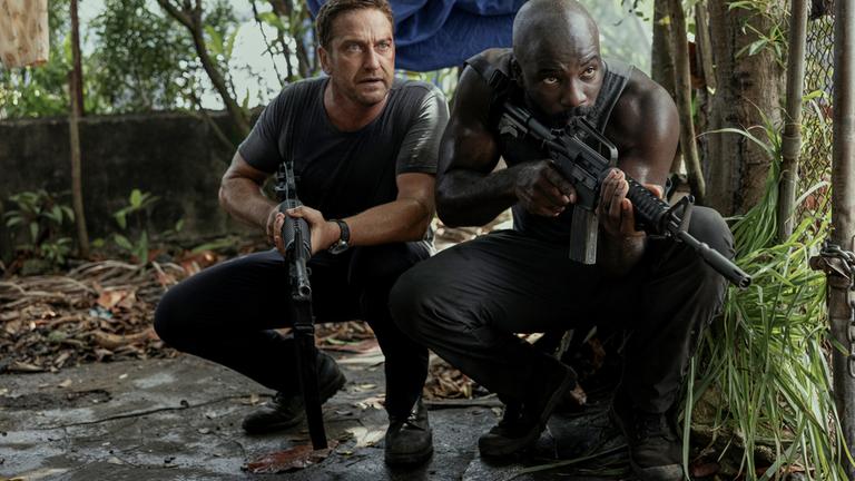 "Plane - Absturz im Dschungel": Brodie Torrance (Gerard Butler) und Louis Gaspare (Mike Colter) hocken auf einem Zementboden im Dschungel und haben ihre Waffen im Anschlag.