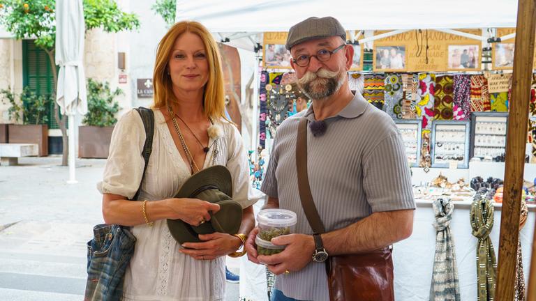 "Horst Lichters Traumrouten - Unterwegs auf Mallorca": Esther Schweins und Horst Lichter stehen vor einem Marktstand und schauen lächelnd in die Kamera. Horst Lichter hält die gemeinsamen Essenseinkäufe in den Händen.