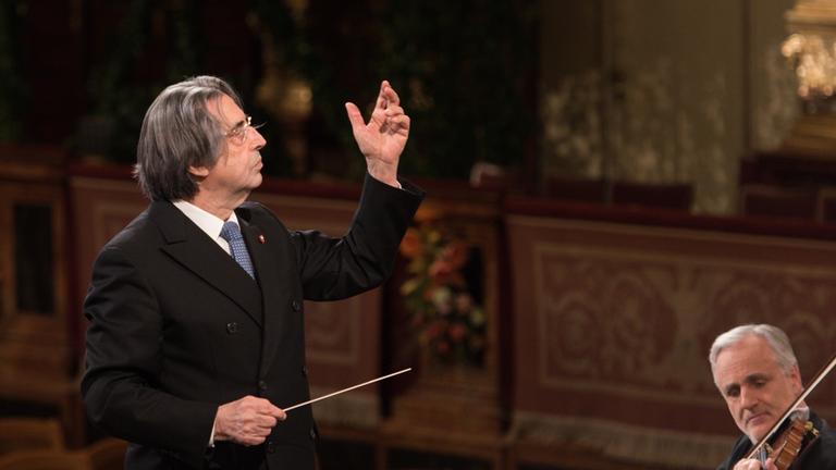 "Neujahrskonzert der Wiener Philharmoniker": Der Dirigent Riccardo Muti steht mit einem erhobenen Arm am Dirigentenpult, in der anderen Hand hält er seinen Dirigentenstab. Er ist im Profil fotografiert, ebenfalls sichtbar im Anschnitt rechts unten ist ein Orchestermitglied an der Geige.