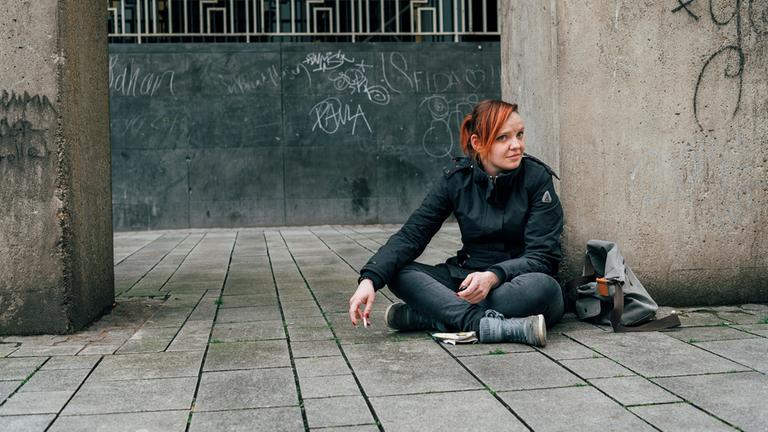 37° "Schutzlos ausgeliefert - Zwischen Straße und Notunterkunft": Leonie sitzt im Schneidersitz auf dem Boden vor einem Betonfeiler. Sie hat rote Haare und trägt schwarze Kleidung. Neben ihr liegt eine Tasche auf dem Boden.