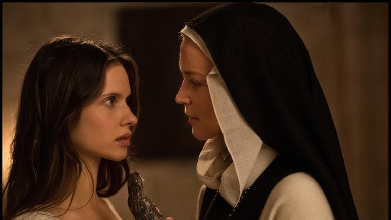 „Benedetta“: Benedetta (Virginie Efira, rechts, im Nonnengewand) und die Novizin Bortolomea (Daphné Patakia, in einem weißen Hemdchen) stehen dicht beeinander und blicken sich intensiv in die Augen. Benedetta hält eine aus Holz geschnitzte Figur in der Hand.
