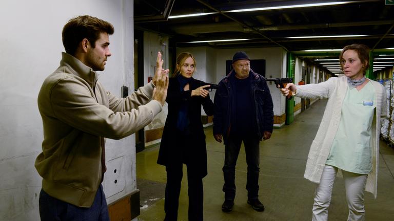 "Ein starkes Team - Der tote Mörder": Marie Lehmann (Jeanette Hain) steht in einem dunklen Kellergang und hat eine Pistole auf Nils Makowski (Lucas Reiber) gerichtet, der mit erhobenen Händen da steht. Linett (Stefanie Stappenbeck) steht ein paar Schritte entfernt zwischen ihnen und hat ihre eigene Waffe auf Marie gerichtet. Neben ihr befindet sich Otto (Florian Martens), der Marie im Blick hält.