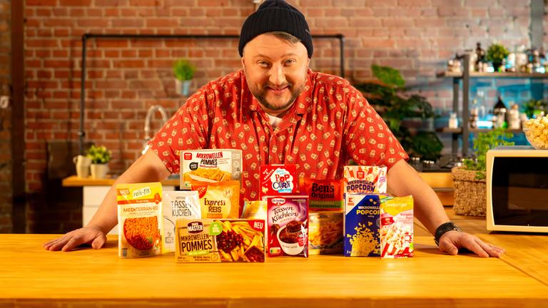 "besseresser: Miese Mikrowellen-Snacks - Sebastian Lege packt aus": Sebastian Lege steht hinter einem Tisch, vor ihm sind verschiedene Mikrowellenprodukte aufgereiht, darunter Popcorn und Reis.