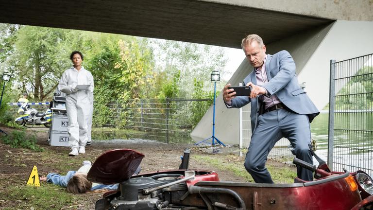 "Marie Brand und das tote Au-pair": Am Ufer neben einer Brücke: Jürgen Simmel (Hinnerk Schönemann) fotografiert mit seinem Handy einen zerstörten Motorroller. Im Hintergrund liegt die Leiche des Au-pairs Alice Taylor (Sonja Joanne Geller). Die Gerichtsmedizinerin Dr. Magda Zimmer (Davina Donaldson) kommt mit einem Klemmbrett in der Hand auf Jürgen Simmel zu.