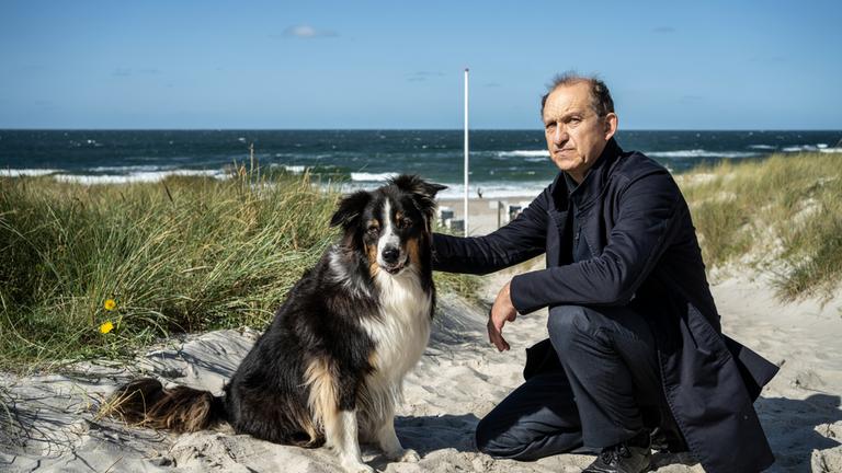 "Nord Nord Mord - Sievers und der verlorene Hund": Carl Sievers (Peter Heinrich Brix) kniet neben einem Australian Shepherd im Sand. Mit seiner rechten Hand streichelt er den Hund, während sein Blick nach vorne geht. Im Hintergrund sind das Gras der Dünen, sowie der Strand und das Meer zu sehen.