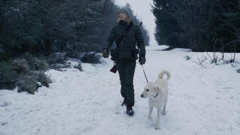 "Erzgebirgskrimi - Wintermord": Die Försterin Saskia Bergelt (Teresa Weißbach) läuft mit Hund Wolke einen zugeschneiten Waldweg entlang. Sie trägt ihr Gewehr auf ihrem Rücken.