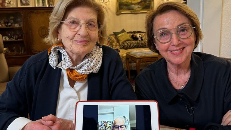 "frontal-Dokumentation: Judenhass in Deutschland - 80 Jahre nach Auschwitz":  Eva Szepesi und Eva Umlauf sitzen lächelnd nebeneinander an einem Tisch. Vor ihnen steht ein Tablet, auf dem Naftali Fürst zu sehen ist