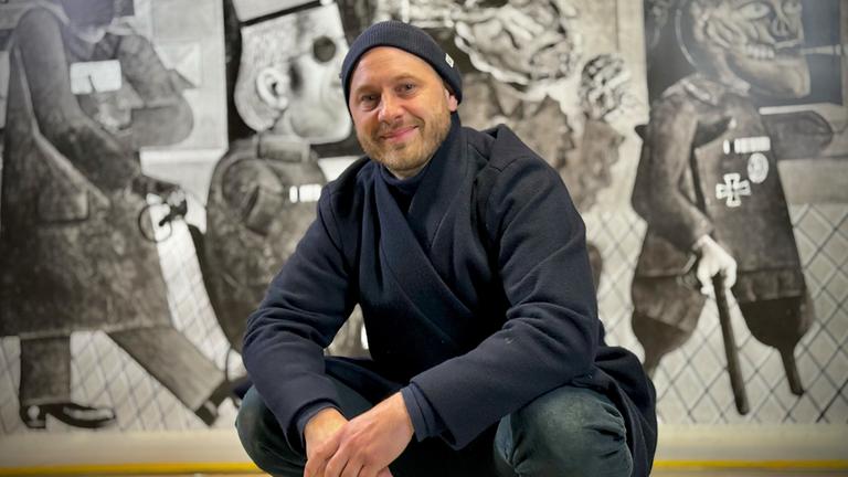"Kunstretter - Im Sturm auf die Moderne": Ein Mann sitzt in Hockstellung vor einem Gemälde und lächelt in die Kamera. Er trägt eine dunkelblaue Mütze und Jacke und eine Jeans. Das Gemälde hinter ihm ist ein großformatiges schwarz-weiß Kunstwerk.