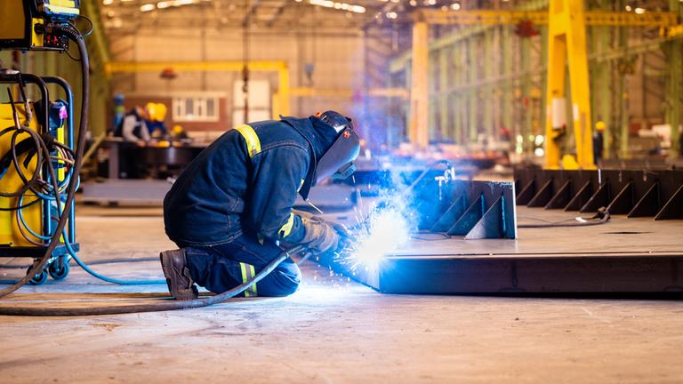 "WISO-Dokumentation: Baustelle Deutschland - Was braucht die Wirtschaft jetzt?": In einer Halle ist ein Arbeiter mit Schweißenarbeiten beschäftigt. Er trägt Schutzkleidung und Schutzmaske. Vor ihm leuchtet das Licht ganz hell und Funken sprühen.