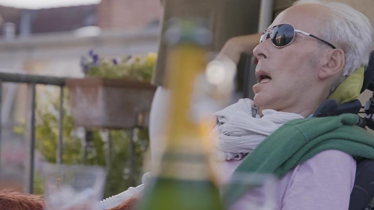 37° Mein Wille geschehe - Wie weit geht die moderne Medizin?": Benedict Mülder liegt auf der Terrasse. Er trägt eine dunkle Sonnenbrille, ein fliederfarbenes Hemd und hat einen grünen Pullover um die Schultern gelegt.