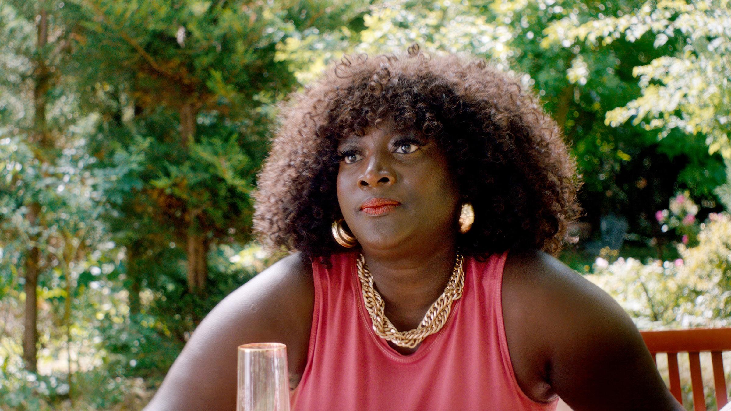 Kisha (Thelma Buabeng) sitzt in einem üppig grünen Garten. Um ihren Hals trägt sie eine massive Goldkette und große goldfarbene Ohrringe.