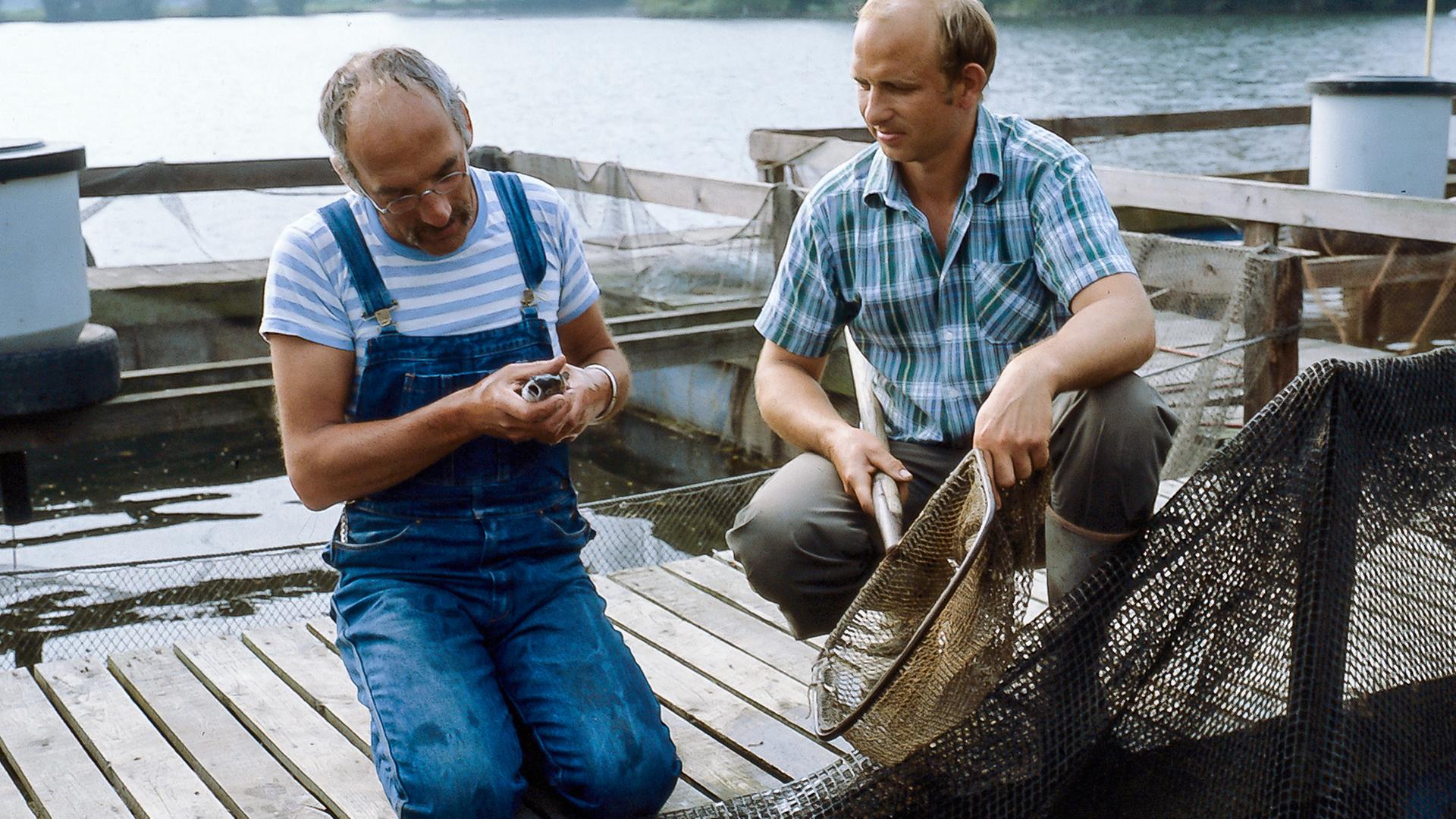 Peter sitzt auf einem Steg am Wasser, neben ihm ein Mann, der einen Kescher hält.